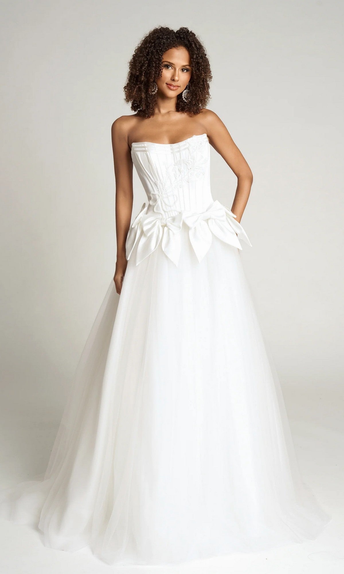 A woman wears the Portia and Scarlett PS26102 strapless white ball gown featuring a fitted corset bodice and large decorative bows at the waist, standing against a plain background.